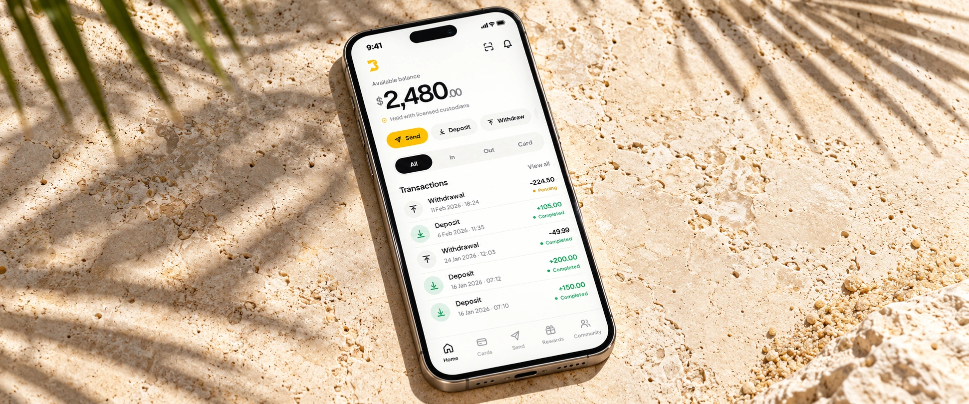 The BullSwipe Home interface on a titanium phone resting on pale coastal stone with palm-leaf shadows.
